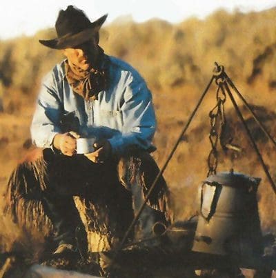 Cowboy drinking coffee outside while sitting beside a campfire with an old iron kettle hung over it.