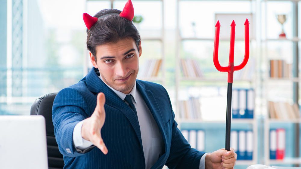 Business man holding plastic red pitchfork and with fake devil horns on his head holds his hand out to shake.