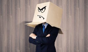 Man in business suit with arms crossed and a cardboard box on his head. The box has a frowning face drawn on it.