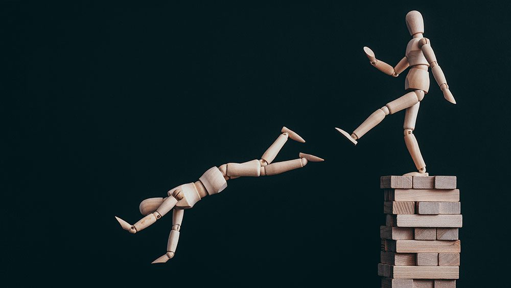 Two wooden mannequins - one kicking the other off a high position on stack of wooden blocks